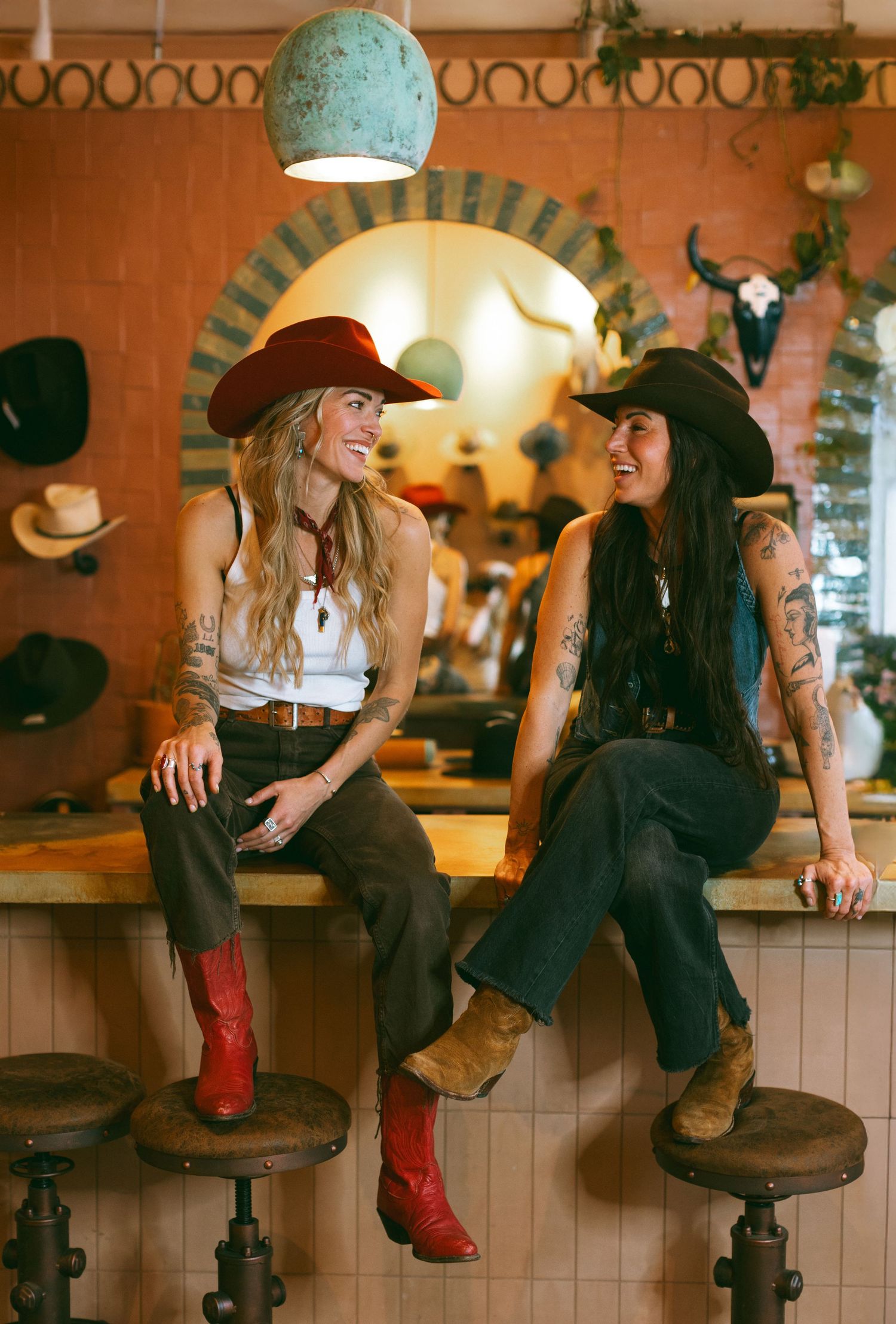 Two fashionable women in western attire and cowboy hats sit on bar stools in a rustic ranch-style setting.