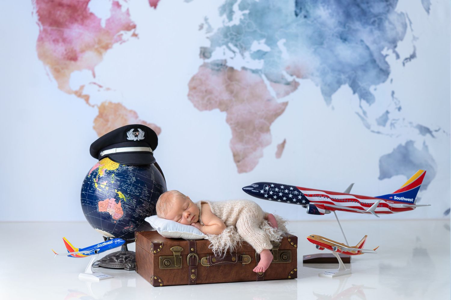 Newborn baby sleeping on vintage suitcase surrounded by toy southwest airplanes, globe, and southwest pilot hat against world map backdrop.