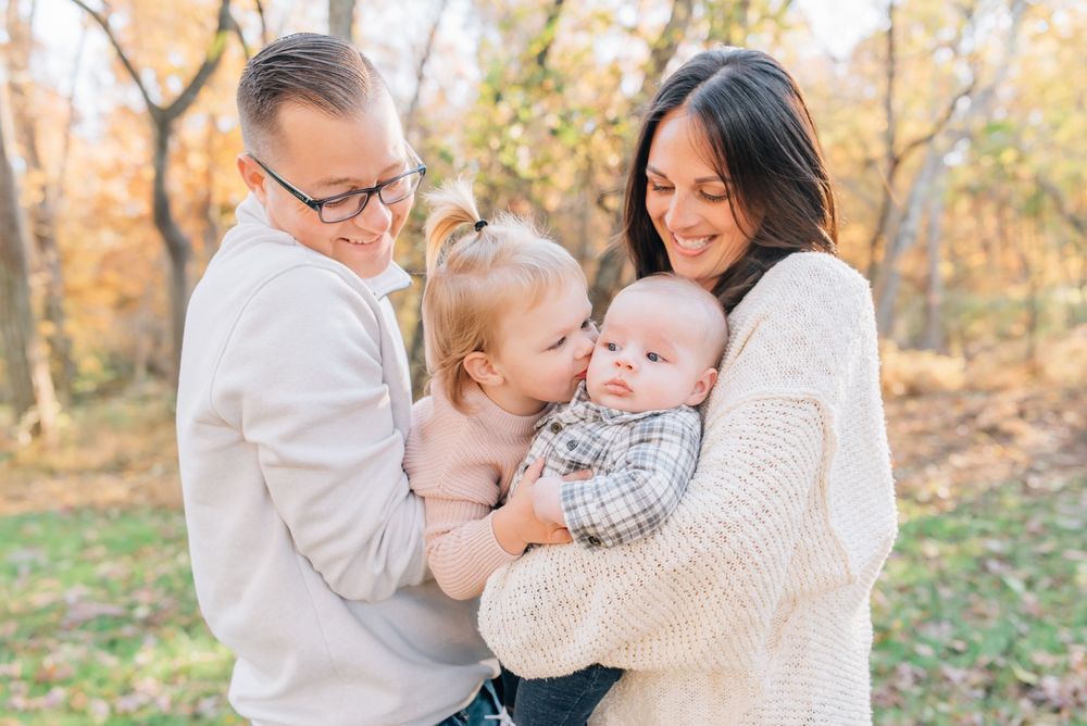 Family of four in fall outfits smiling near historic village buildings at Allaire State Park, NJ.