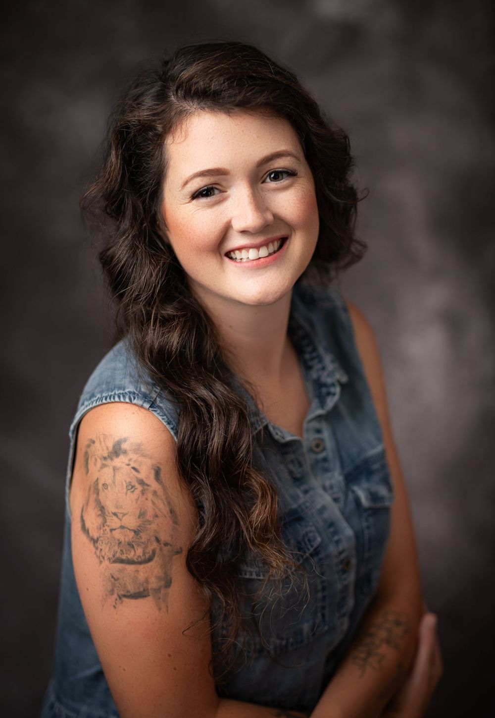 Portrait of a smiling person wearing a denim vest with visible arm tattoo against dark studio background.
