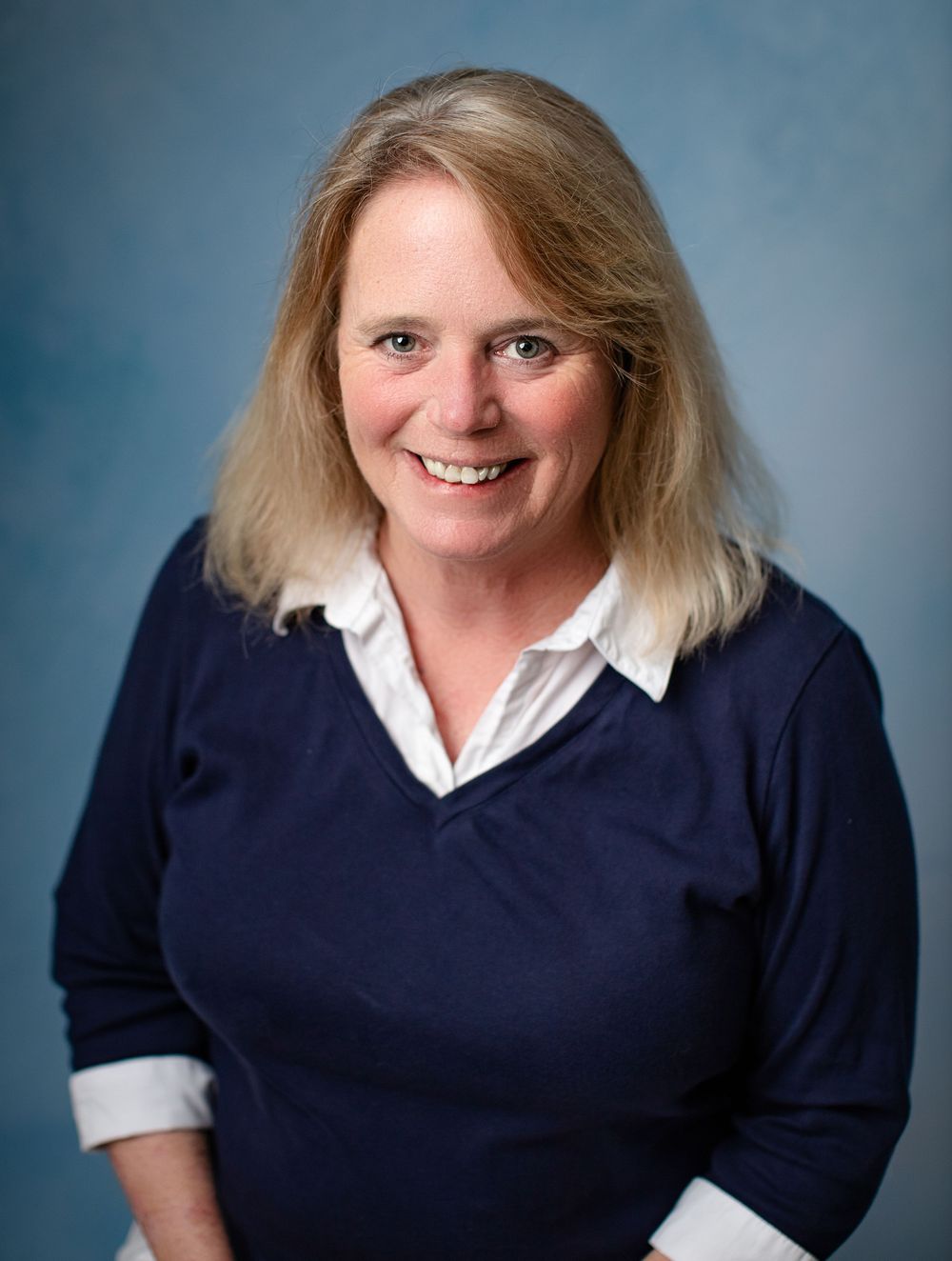 Professional studio portrait photograph with a person wearing a navy blue sweater and white collared shirt against a blue background.