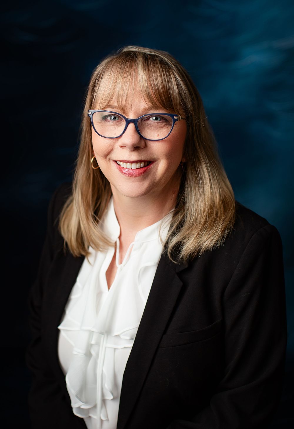 Professional business portrait against blue backdrop showing a person wearing glasses, black blazer and white blouse.