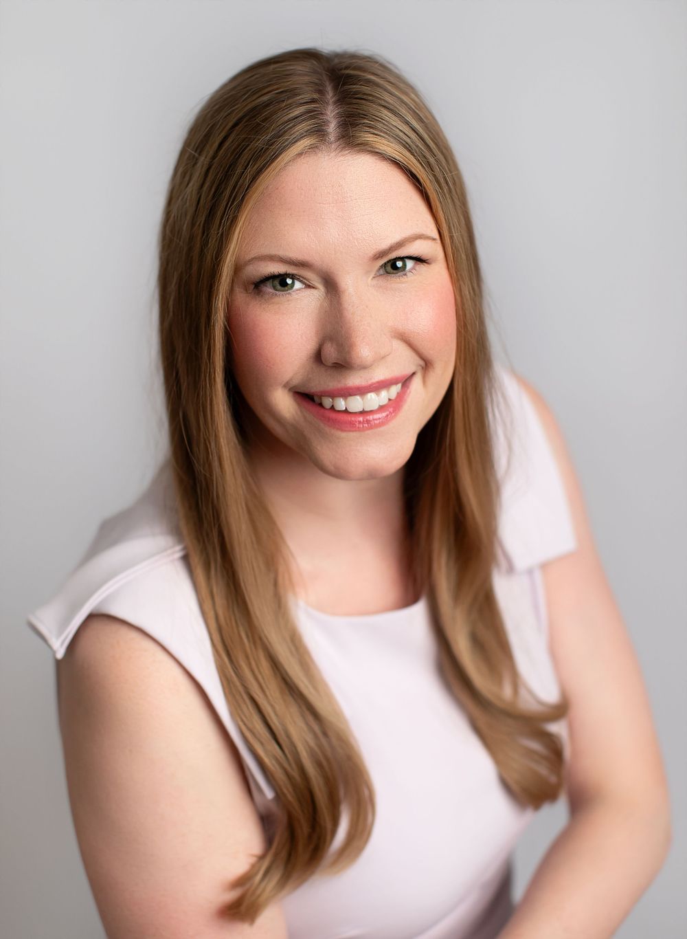 Professional headshot portrait of someone smiling warmly while wearing a white sleeveless top against a neutral gray background.