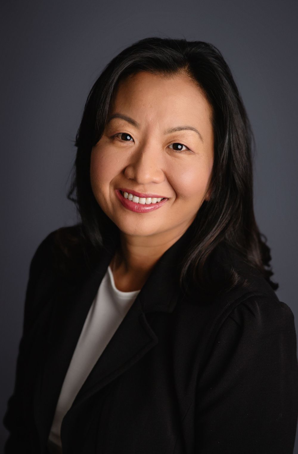 Professional headshot portrait of someone wearing a black blazer and white top against a dark background, smiling warmly.