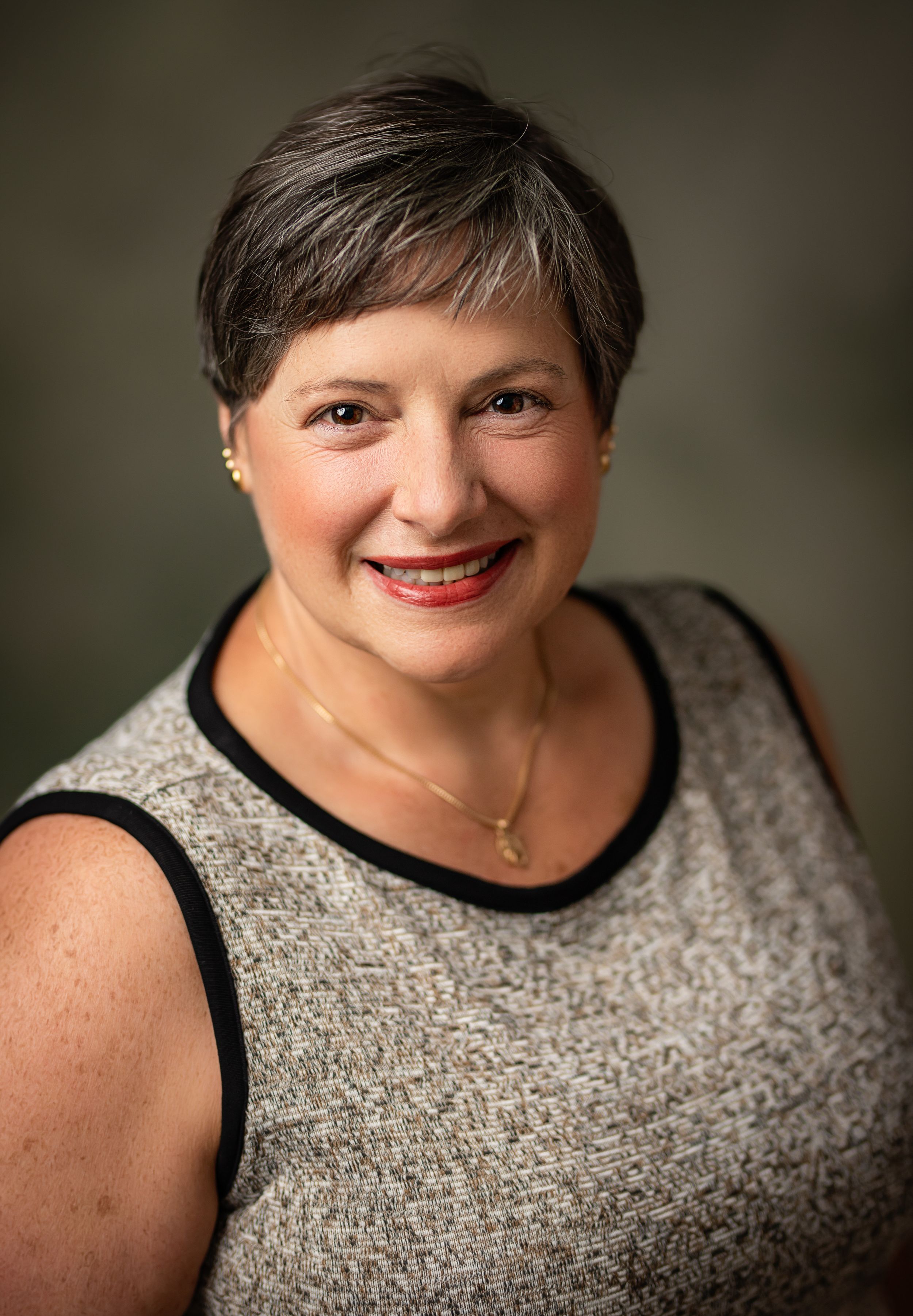 Professional portrait photograph showing someone in a sleeveless sparkly top with a necklace against a dark background.