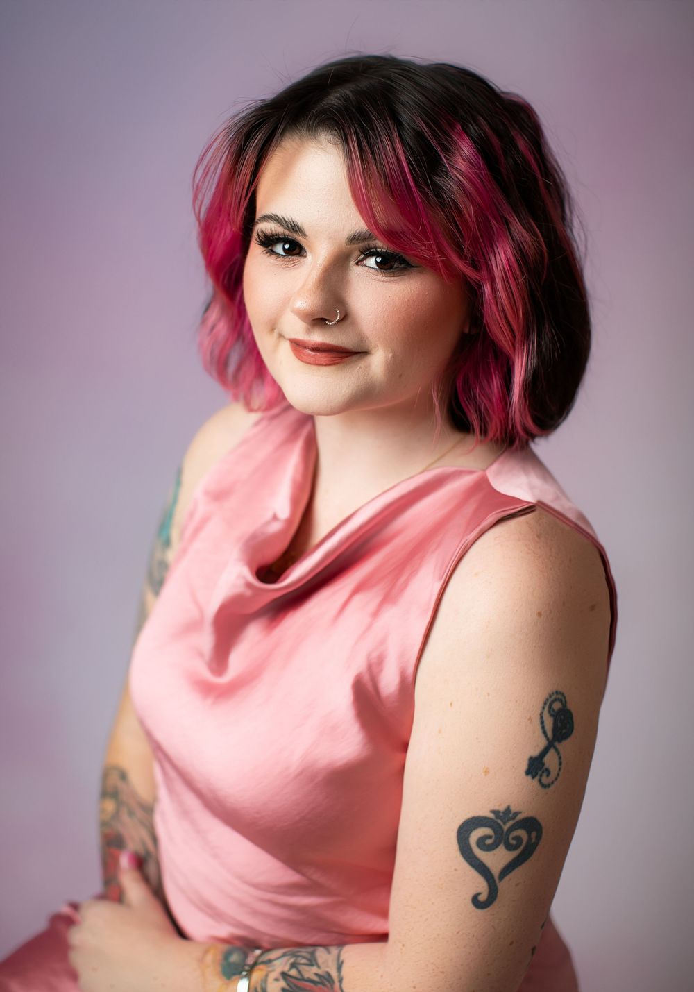 Portrait of someone wearing a pink sleeveless top with dyed pink hair and visible tattoos against a purple background.