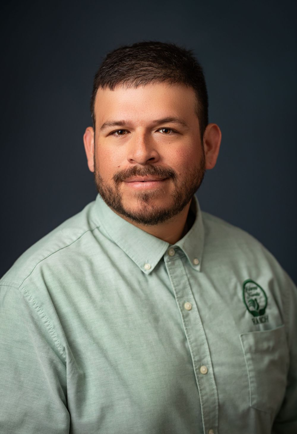 Professional headshot portrait of a person in a sage green button-down shirt against a dark studio background.