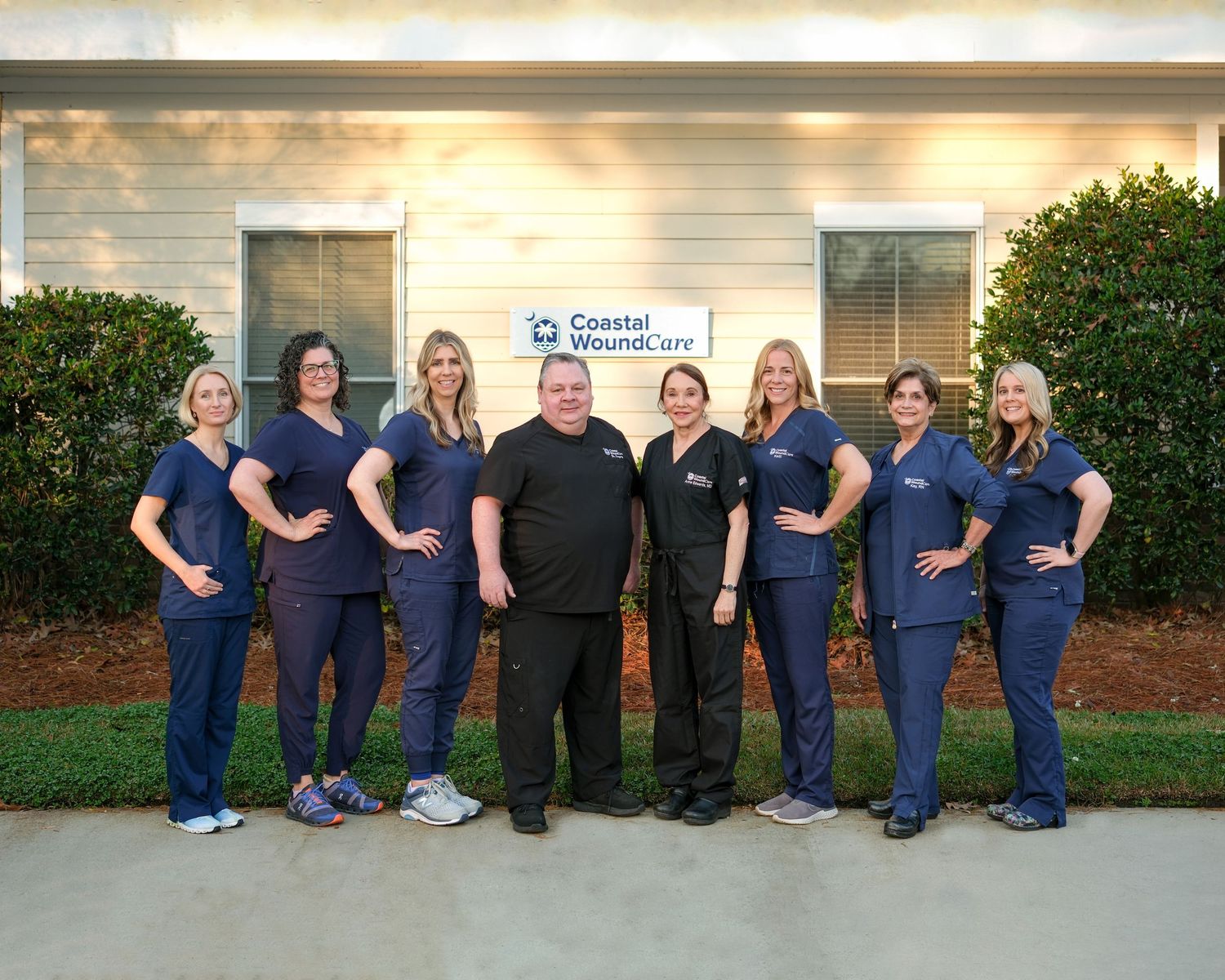 Six medical professionals in matching navy blue scrubs pose together outside a healthcare facility.