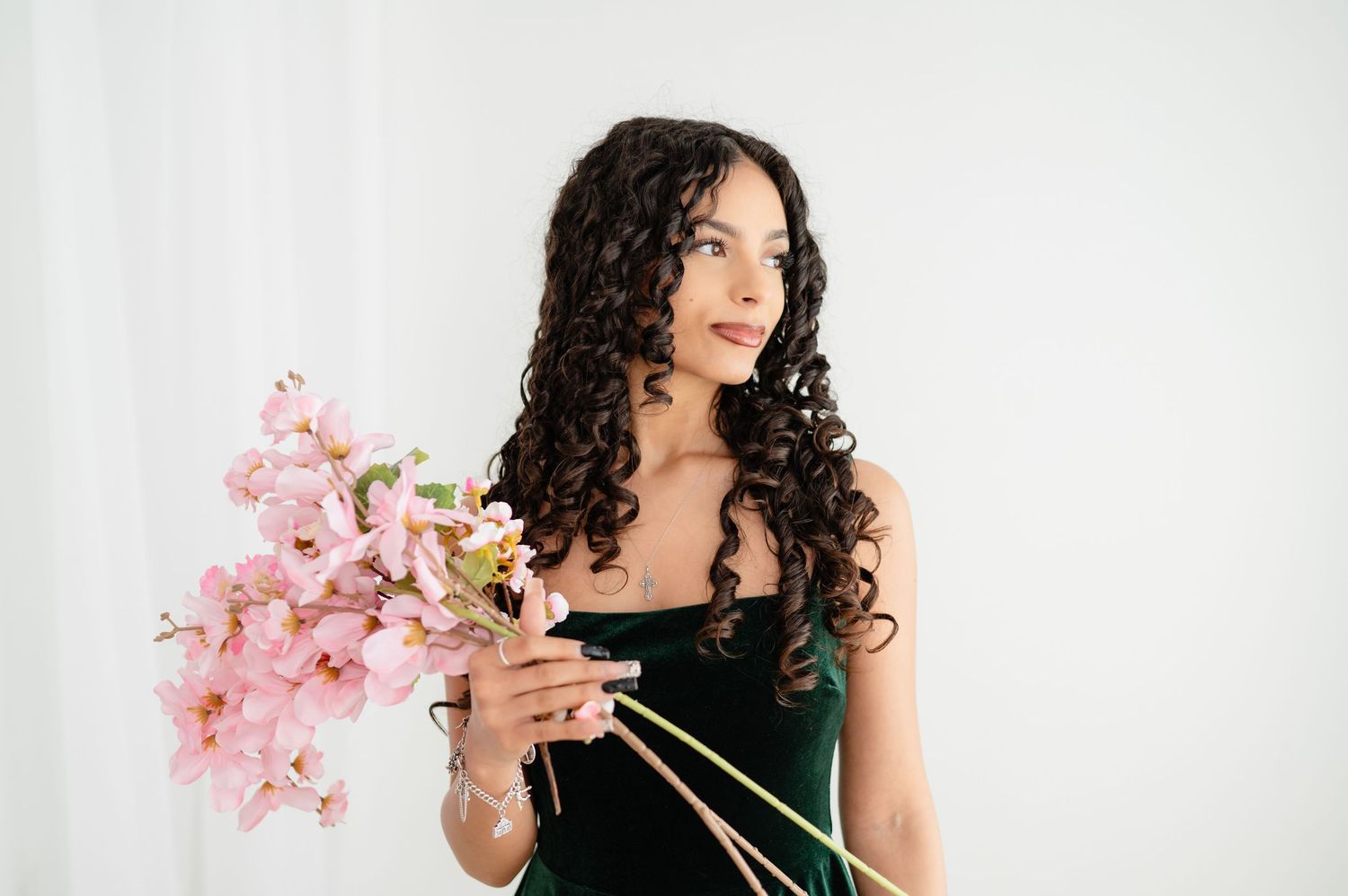 A person with long curly dark hair wearing a black dress holds pink orchid stems against a white background.