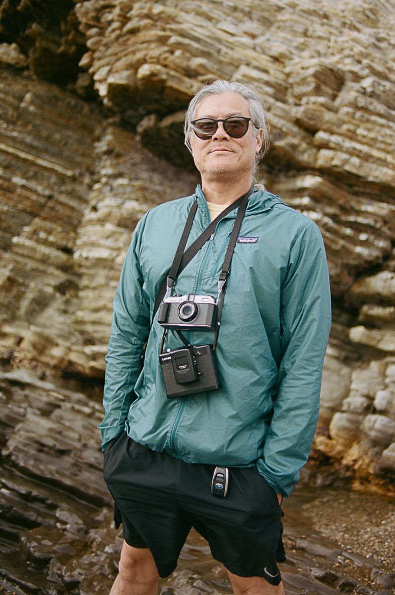 Adventurer wearing sunglasses and green jacket with vintage cameras around neck against rocky cliff backdrop.