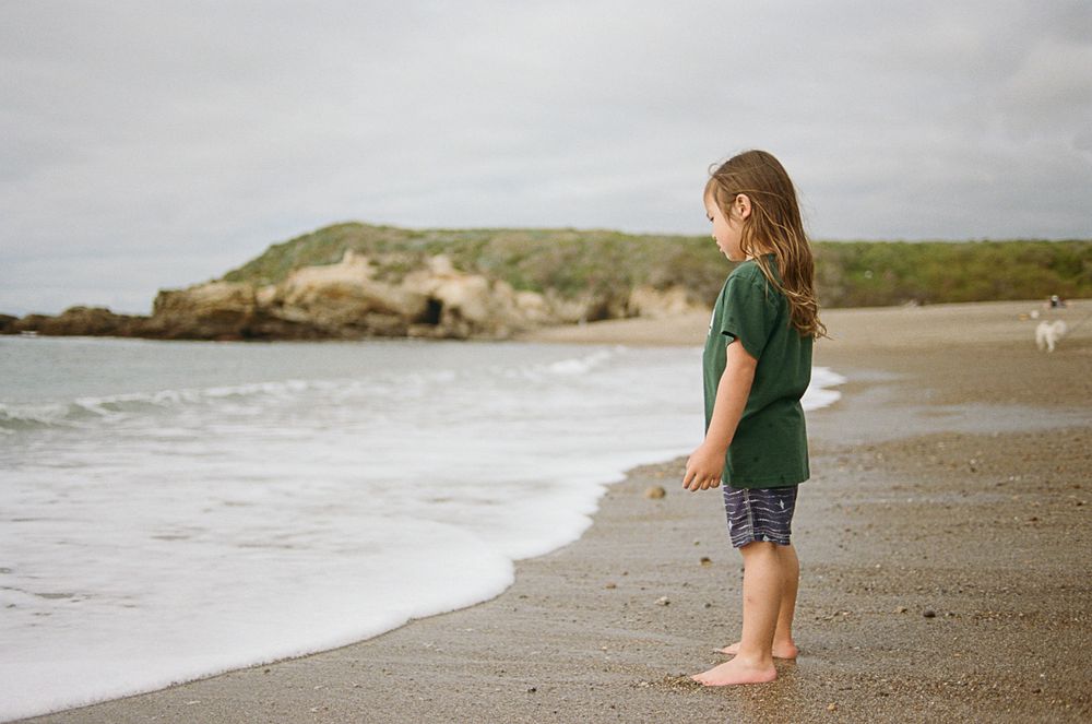 Child standing at water's edge on beach with coastal cliffs in background.