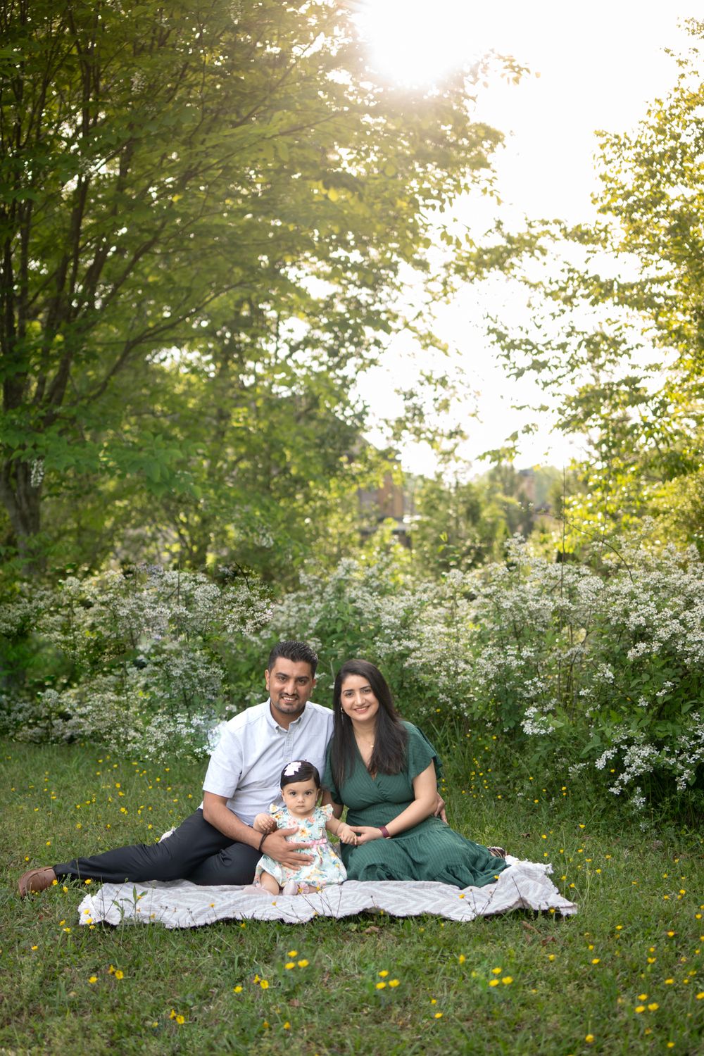 A mom and dad with their 11 month old daughter sitting on a blanket in the grass