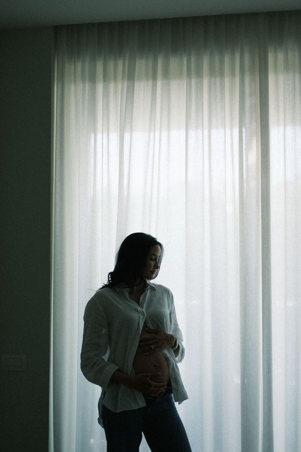 Pregnant woman in white shirt cradling her baby bump while standing in front of sheer curtains.
