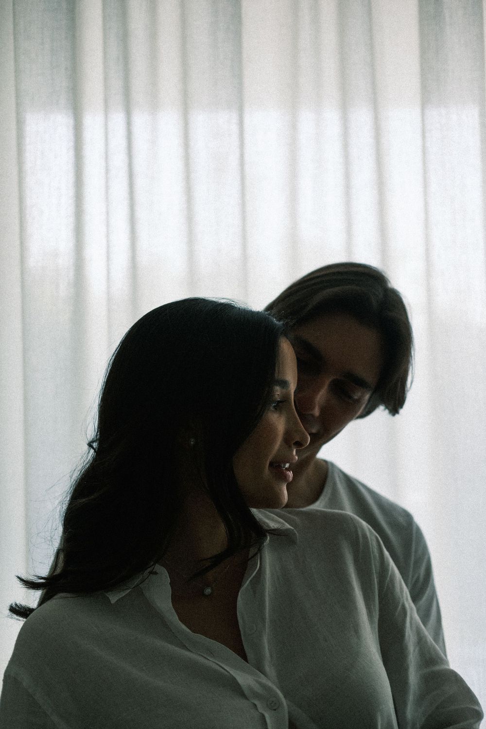 Intimate couple in white linen shirts standing close together near sheer curtains in soft natural light.