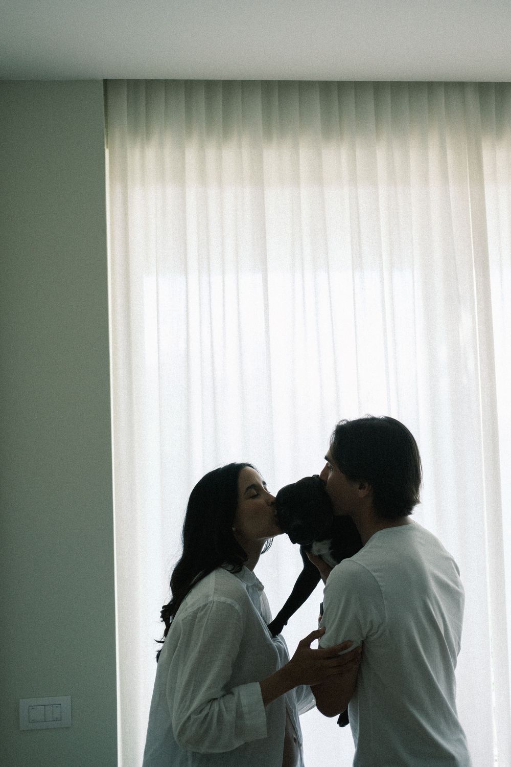Couple sharing a tender kiss while holding a small dog near a bright window with white curtains.
