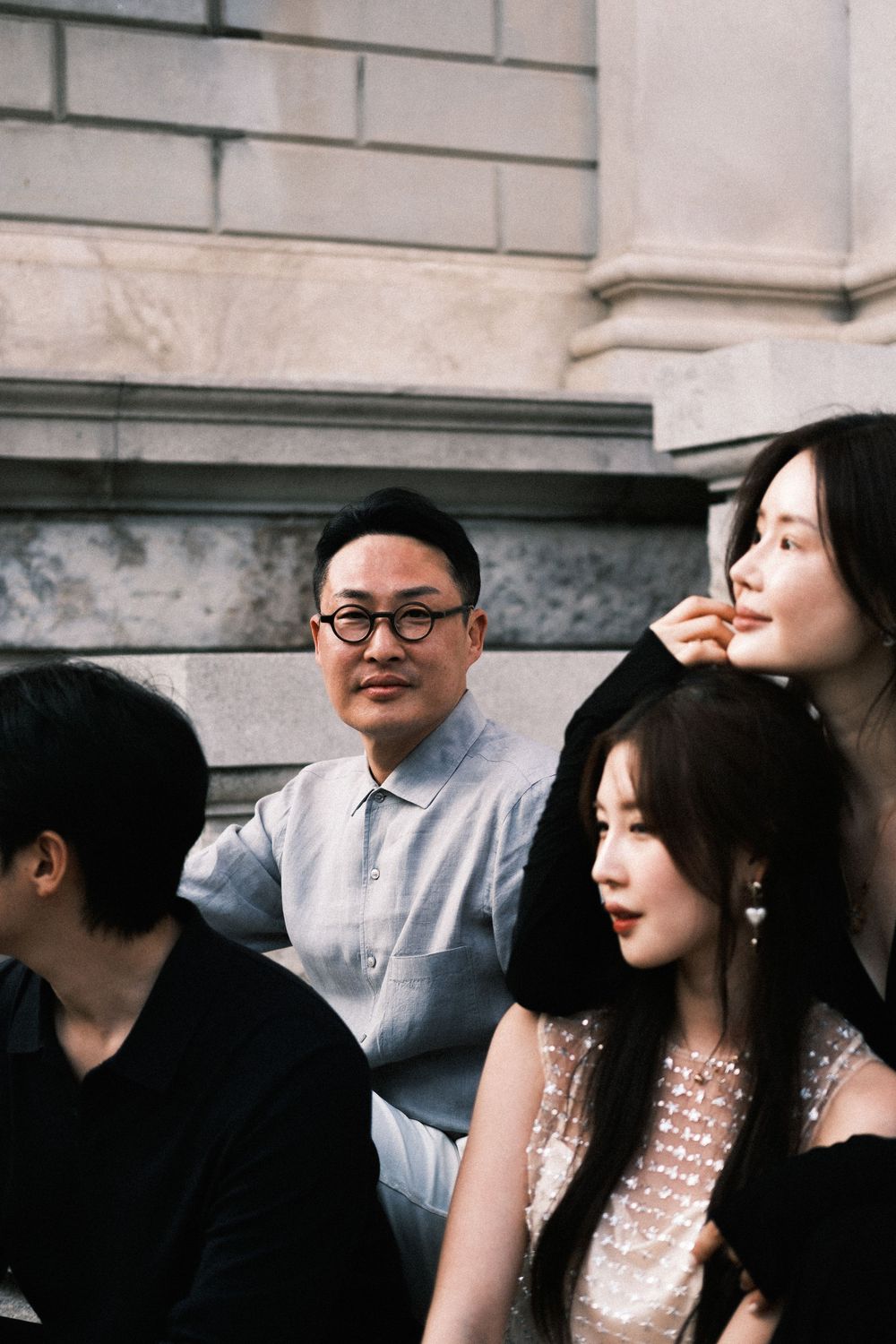 Group of elegantly dressed people sitting on stone steps, man with round glasses in light blue shirt among stylish women.