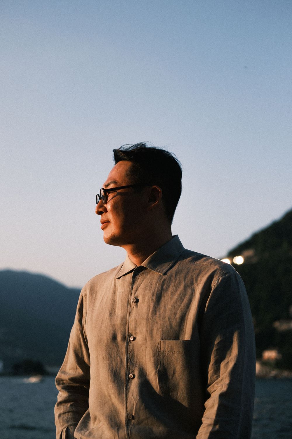 Man in linen shirt and sunglasses standing outdoors at dusk with mountains and water in background.