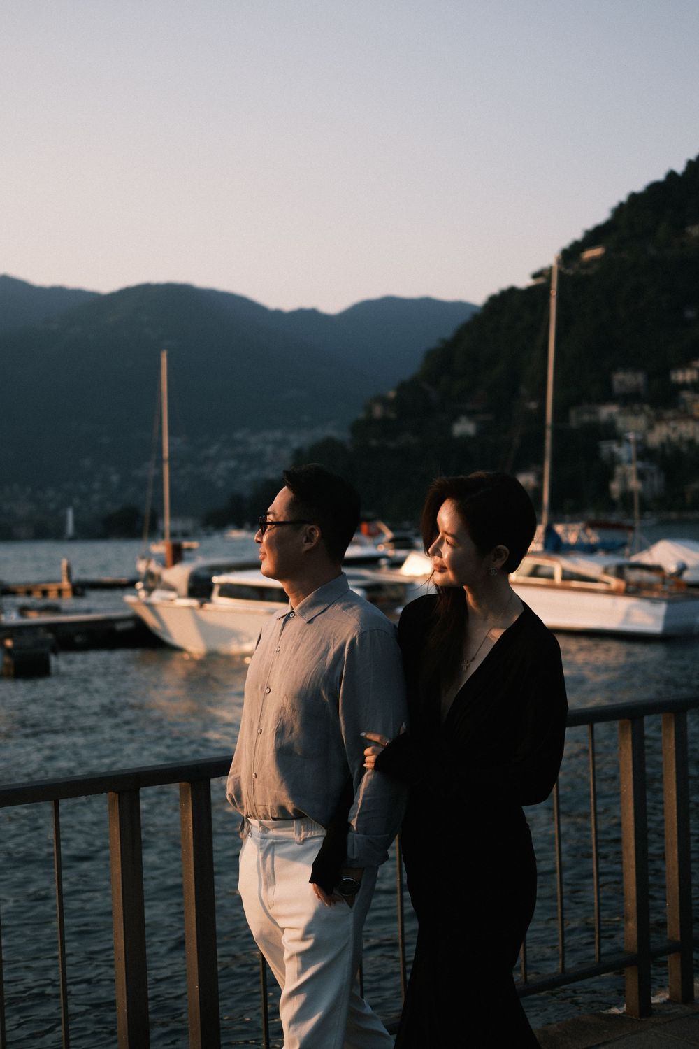 Couple holding hands at sunset by lake marina with sailboats and mountains in background.