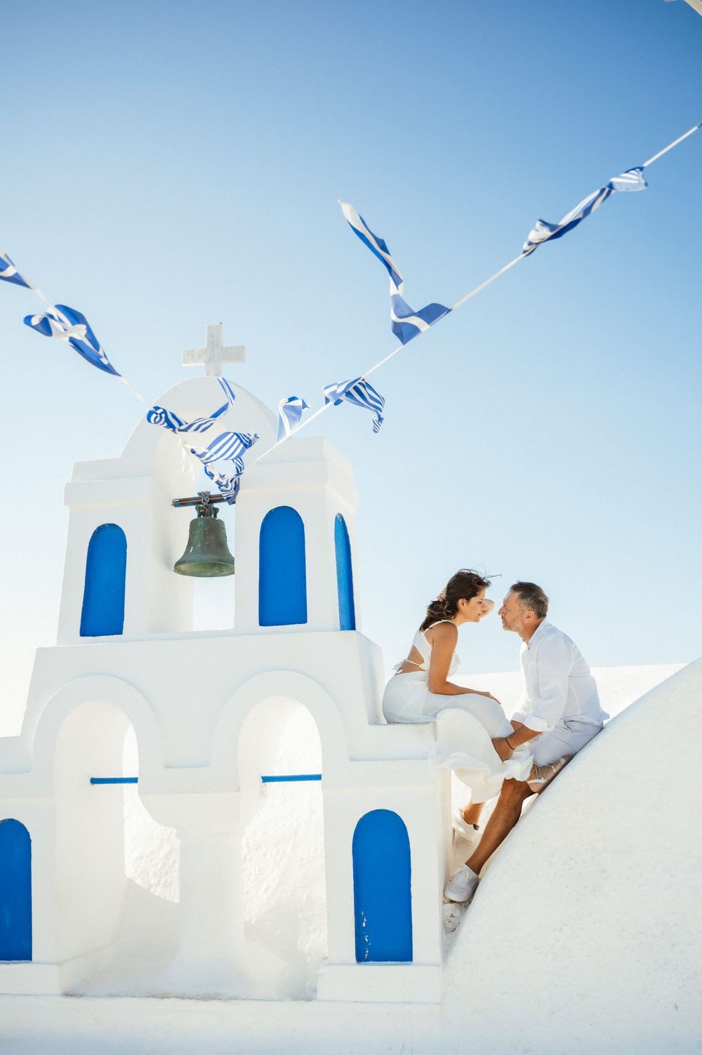 Couple poses on white Greek church steps with blue doors and fluttering flags overhead.