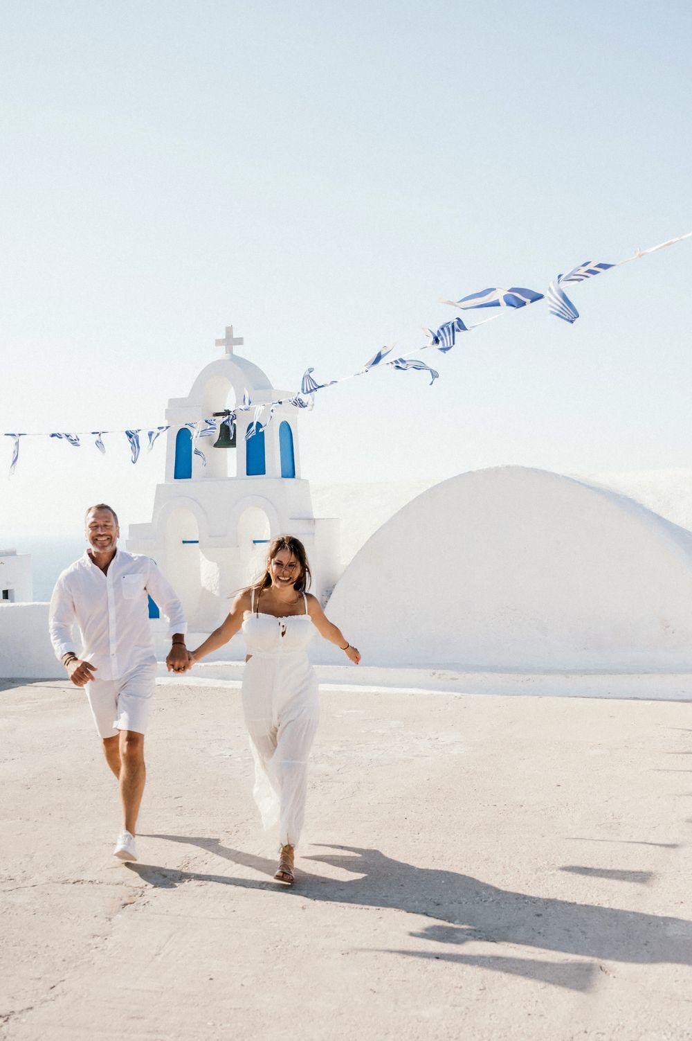 Couple running together in white outfits by traditional Greek church with blue flags.