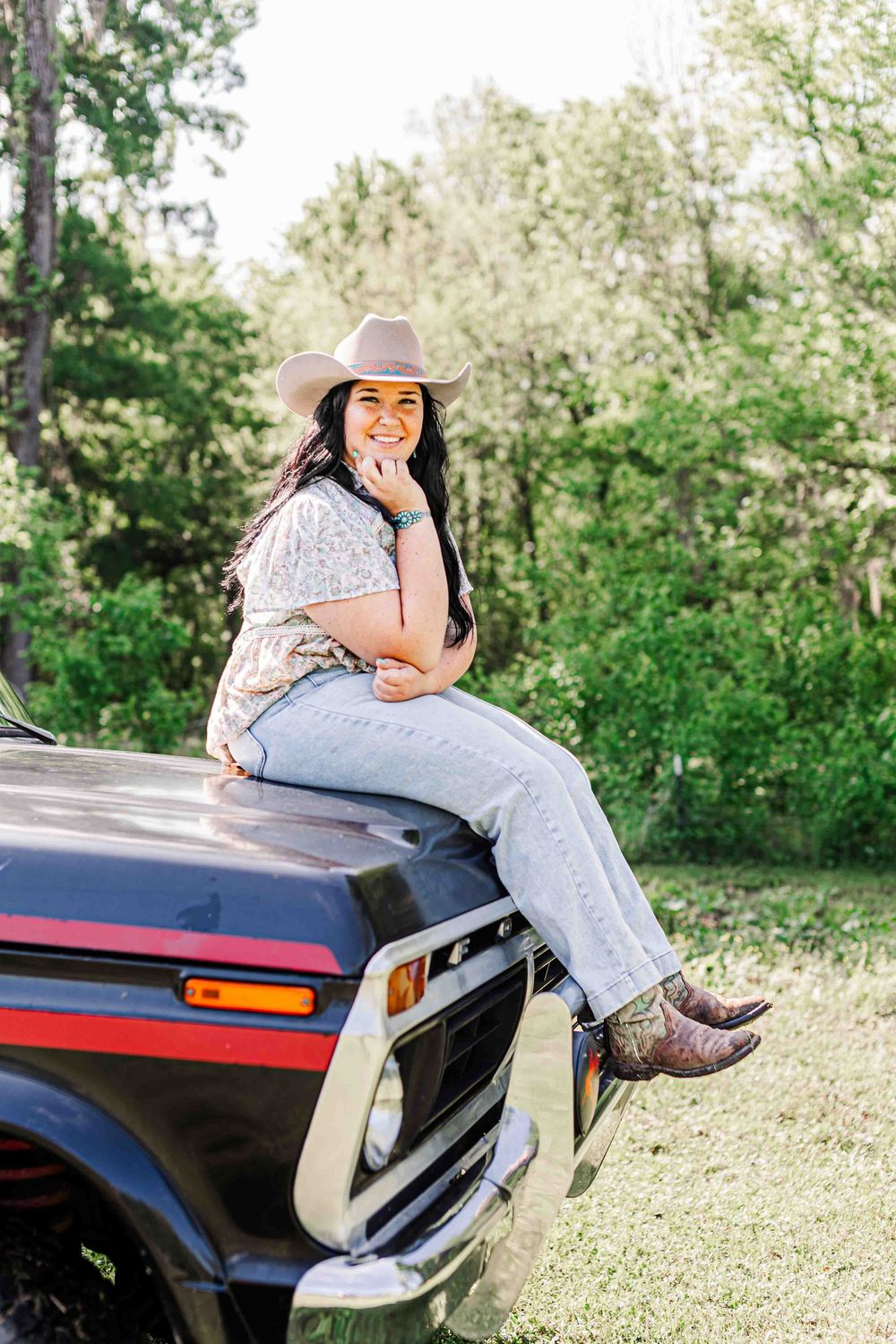A casual country style photo showing someone in a cowboy hat and jeans sitting on a vintage car outdoors.
