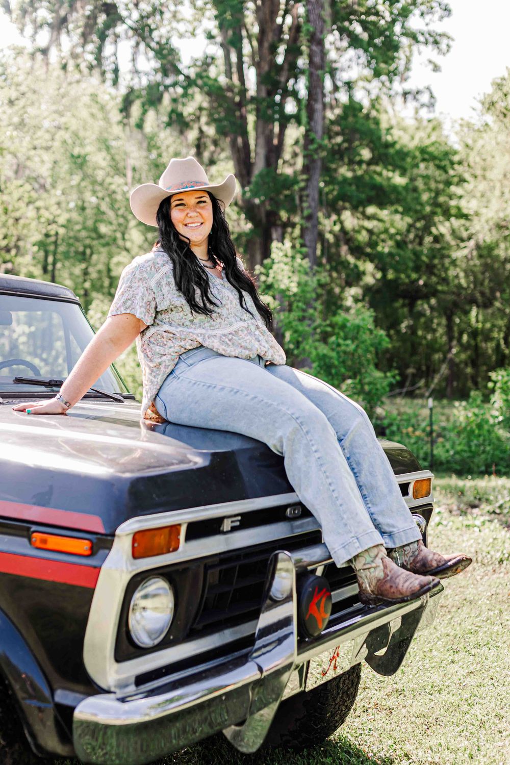 Someone in a cowboy hat and jeans sits casually on the hood of a vintage Ford pickup truck in a sunny outdoor setting.