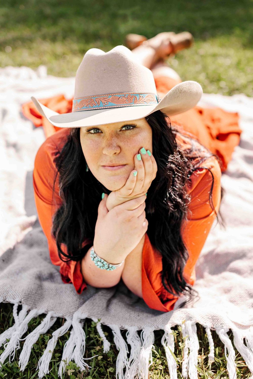 Woman wearing orange top and beige cowboy hat lying on blanket with turquoise jewelry in outdoor setting.