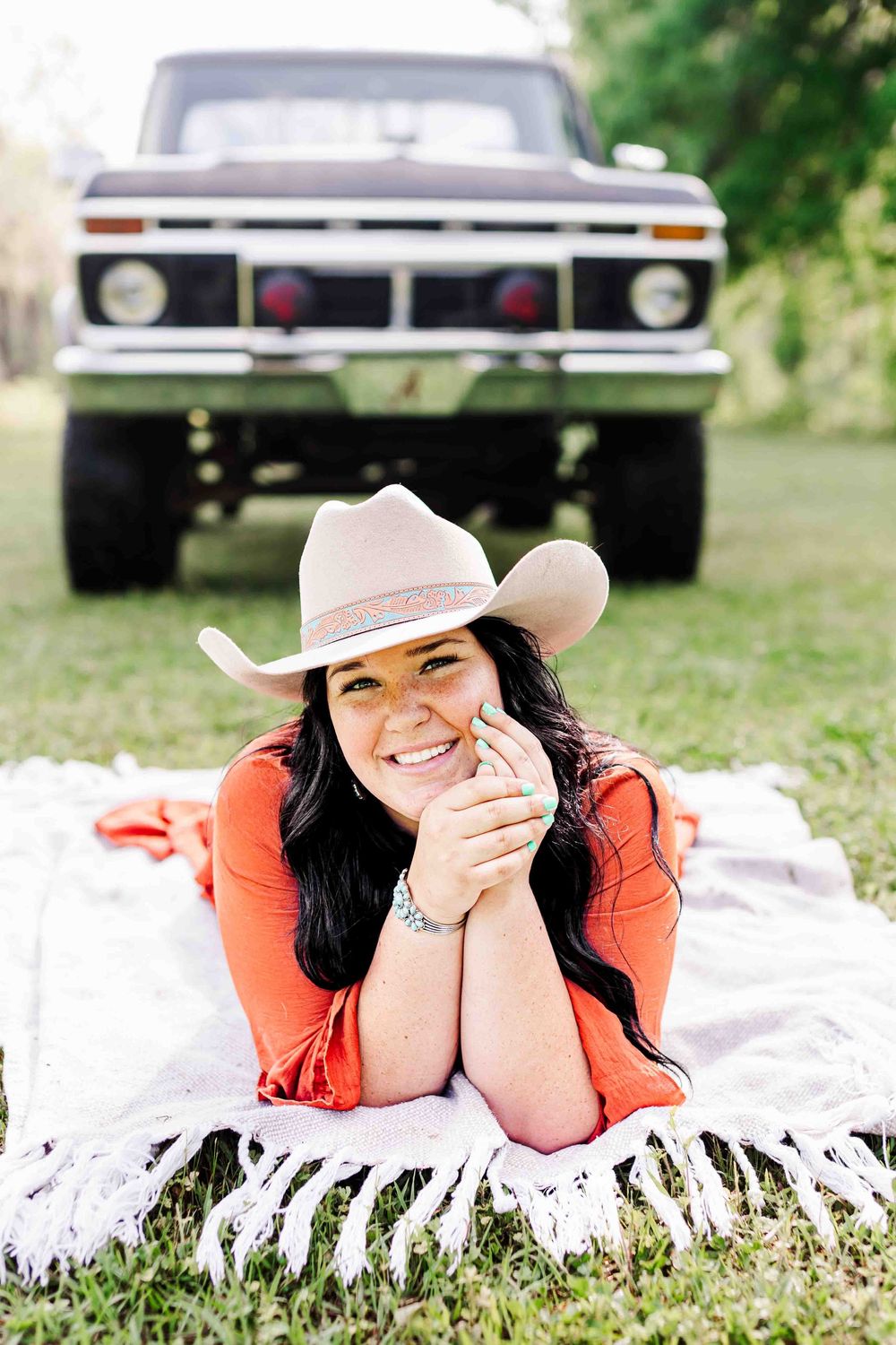 Someone in an orange shirt and light cowboy hat relaxes on a blanket with a vintage Ford pickup truck in the background.