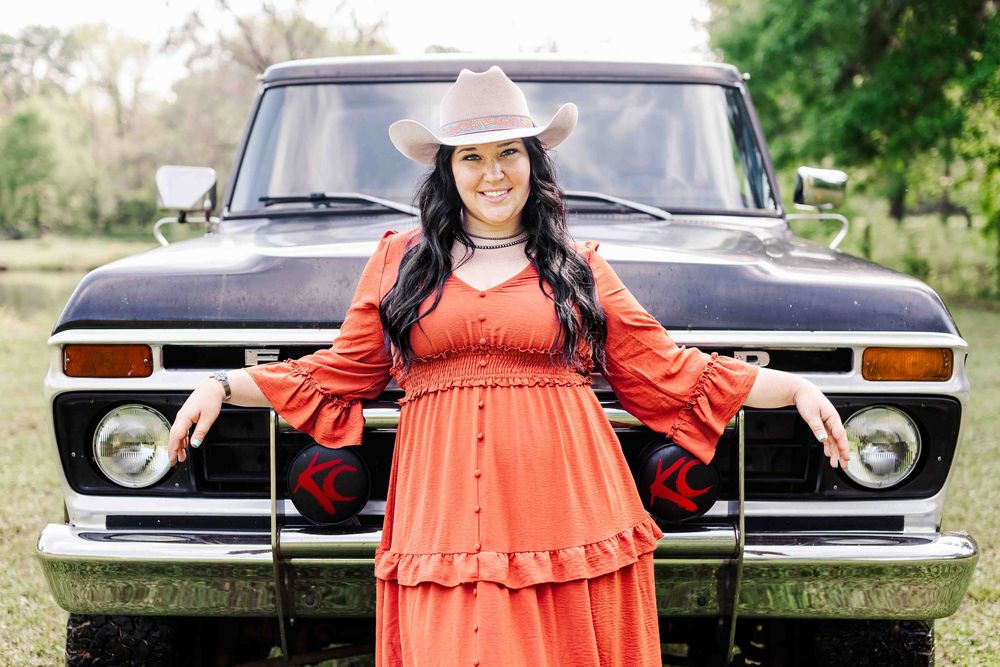 Person wearing an orange dress and cowboy hat poses in front of a vintage Ford pickup truck in a rustic outdoor setting.