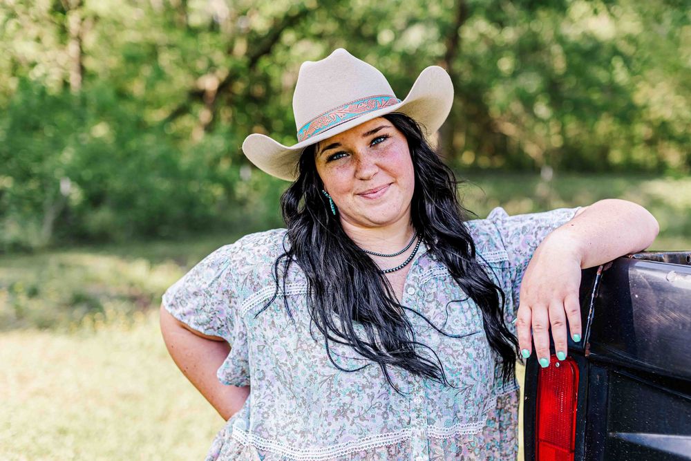 Someone in a cowboy hat and floral top leans casually against a red pickup truck in an outdoor country setting.