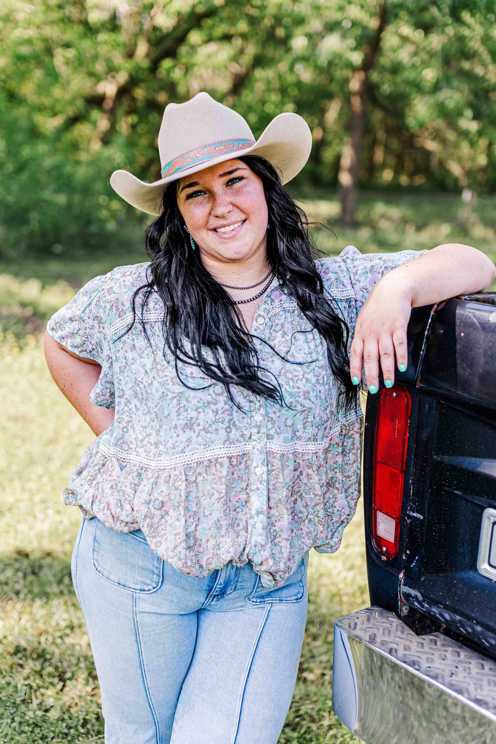 A woman in a cowboy hat and floral top leans against a pickup truck on a sunny day in a rural setting.