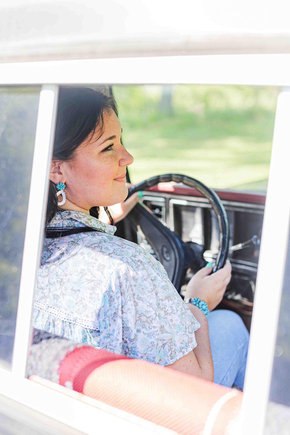 Driver in a light blue patterned shirt and turquoise jewelry sits at the wheel of a vintage car on a sunny day.