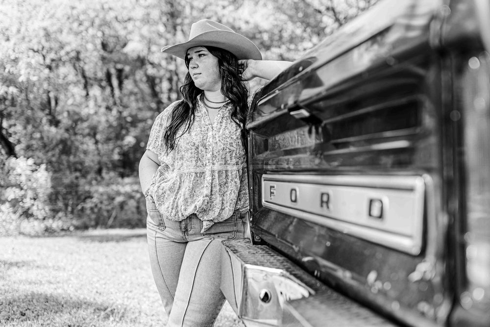 Black and white portrait of a cowgirl in western attire leaning against a vintage Ford truck in a rustic setting.