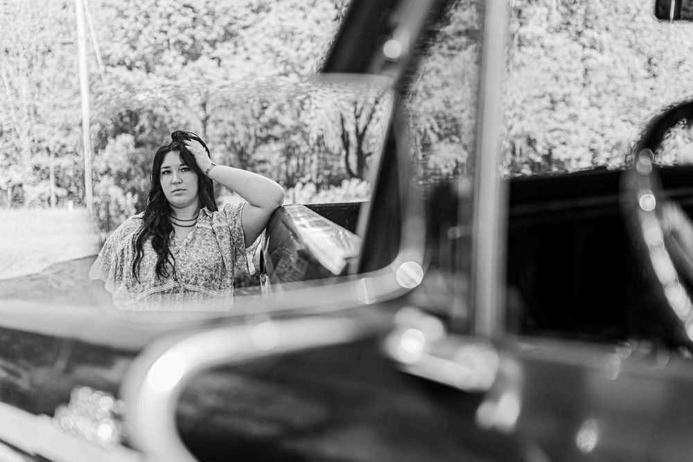 Black and white image of someone in a vehicle with long dark hair, seen through a car window reflection.