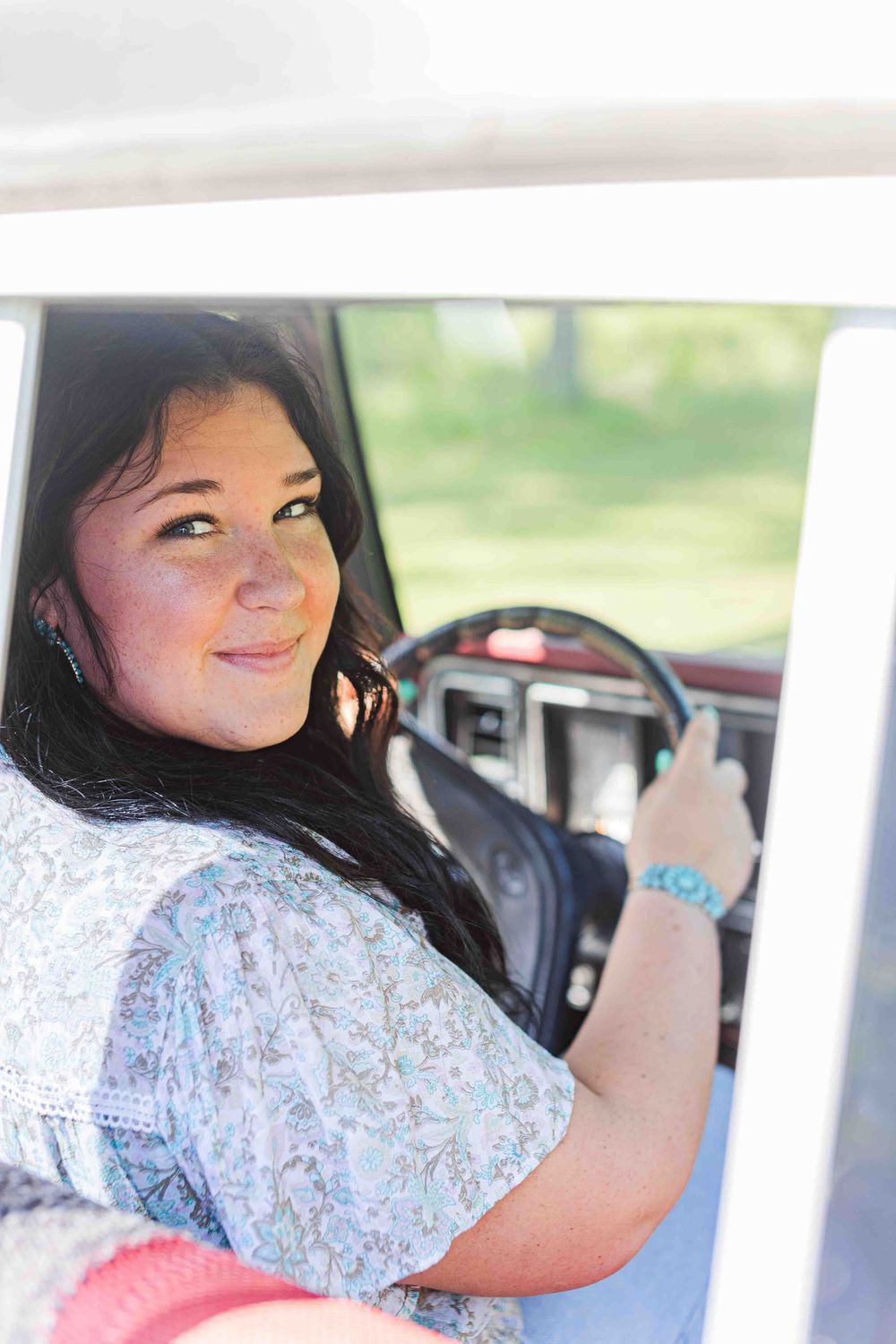 A young woman in a floral print top smiles while sitting in a car holding the steering wheel.
