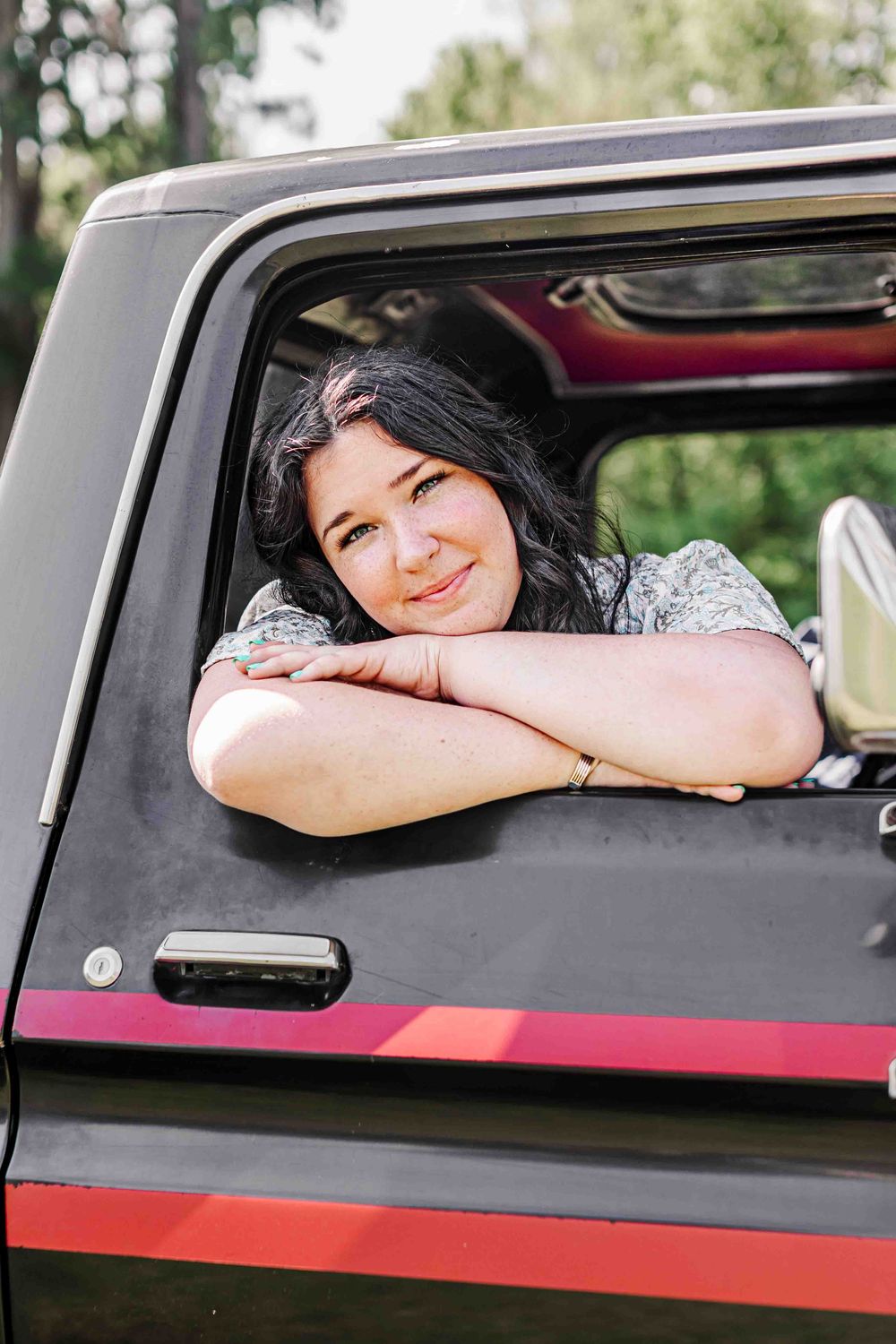 Someone leans on their arm and smiles through the window of a black pickup truck with red striping.
