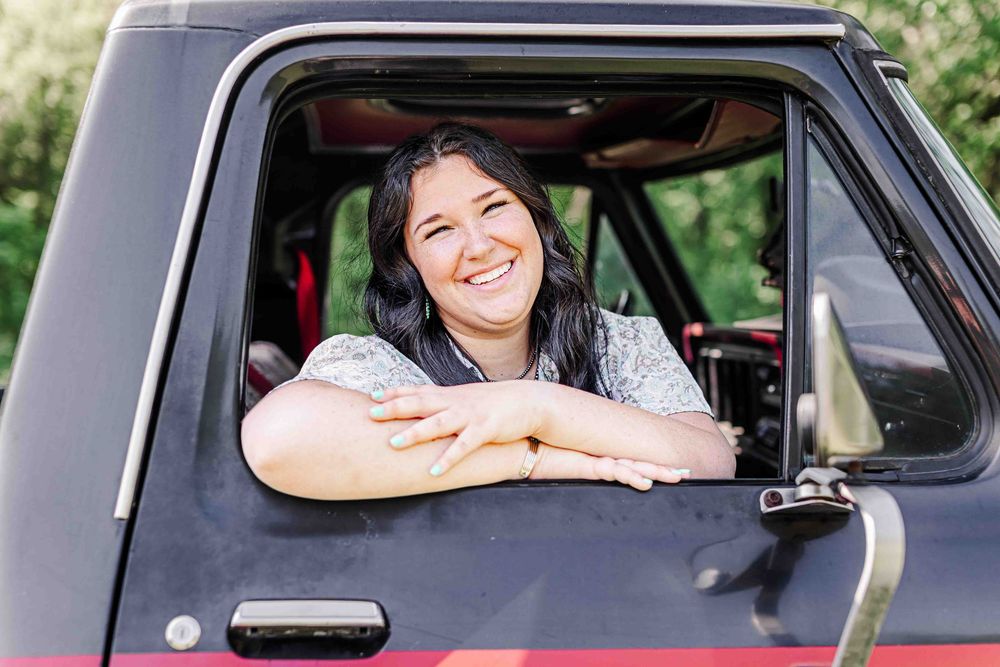 Someone sitting in a vintage black truck and leaning out the driver's window with an expansive smile while outdoors.