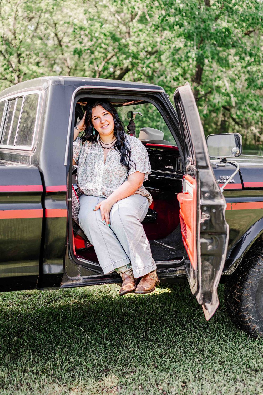 Someone sits casually in the doorway of a black pickup truck wearing a light colored outfit on a sunny day.