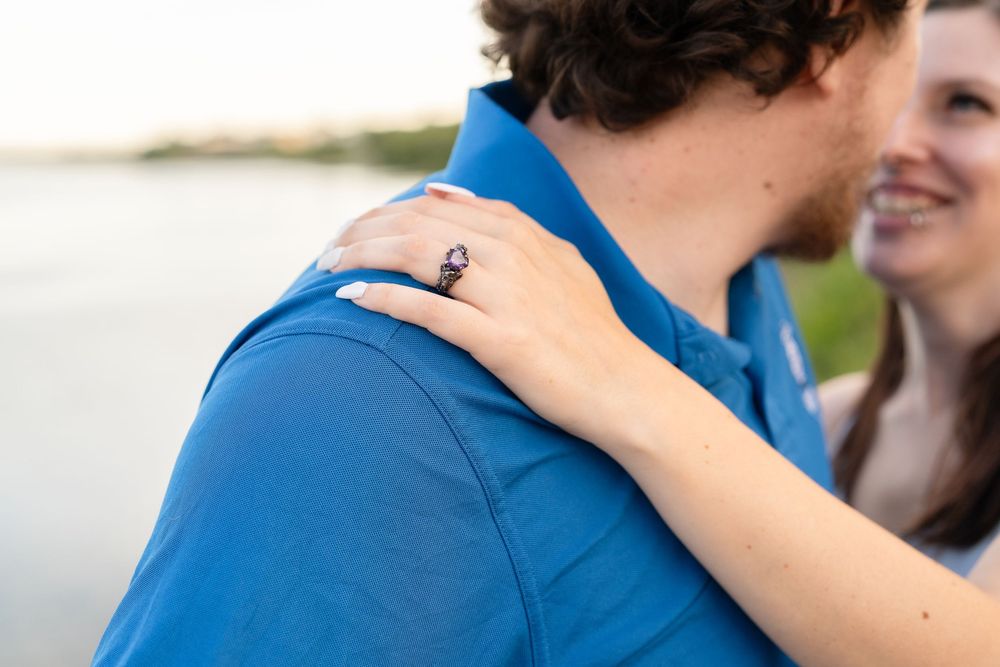 A hand wearing an engagement ring rests on a shoulder of someone in a bright blue shirt near a waterfront.