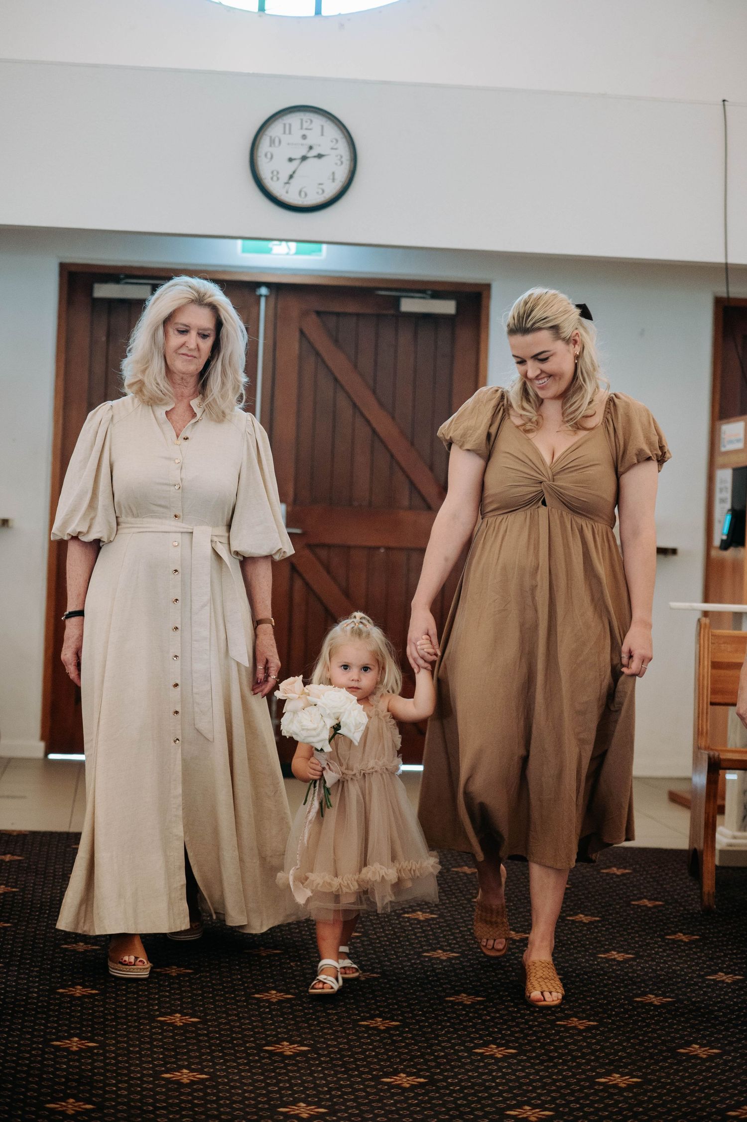 Three people holding hands in vintage-style dresses in neutral earth tones stand together in an indoor space.
