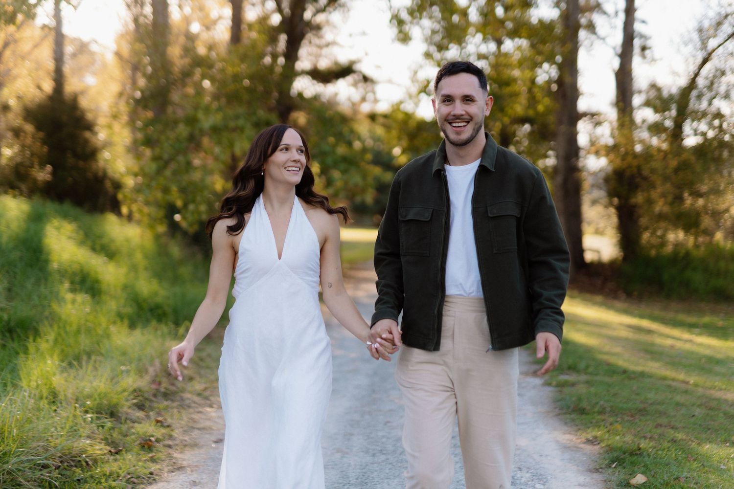 A laughing groom in a navy suit stands with a bride in white dress and veil outdoors at a garden venue.