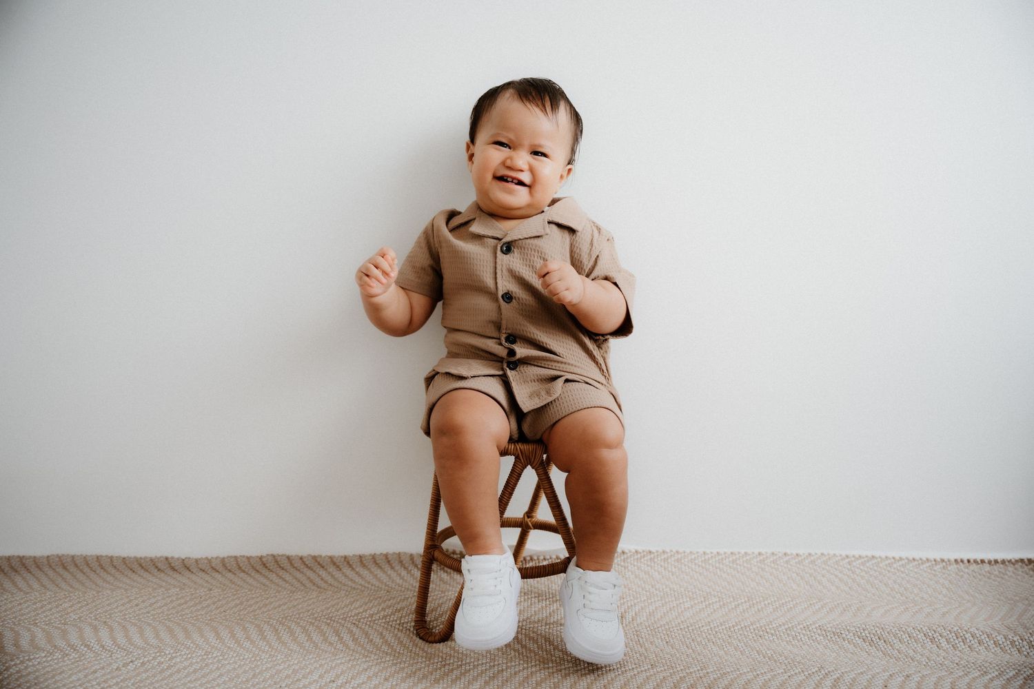 Baby in tan romper outfit sits in small wooden chair while smiling against white wall background.