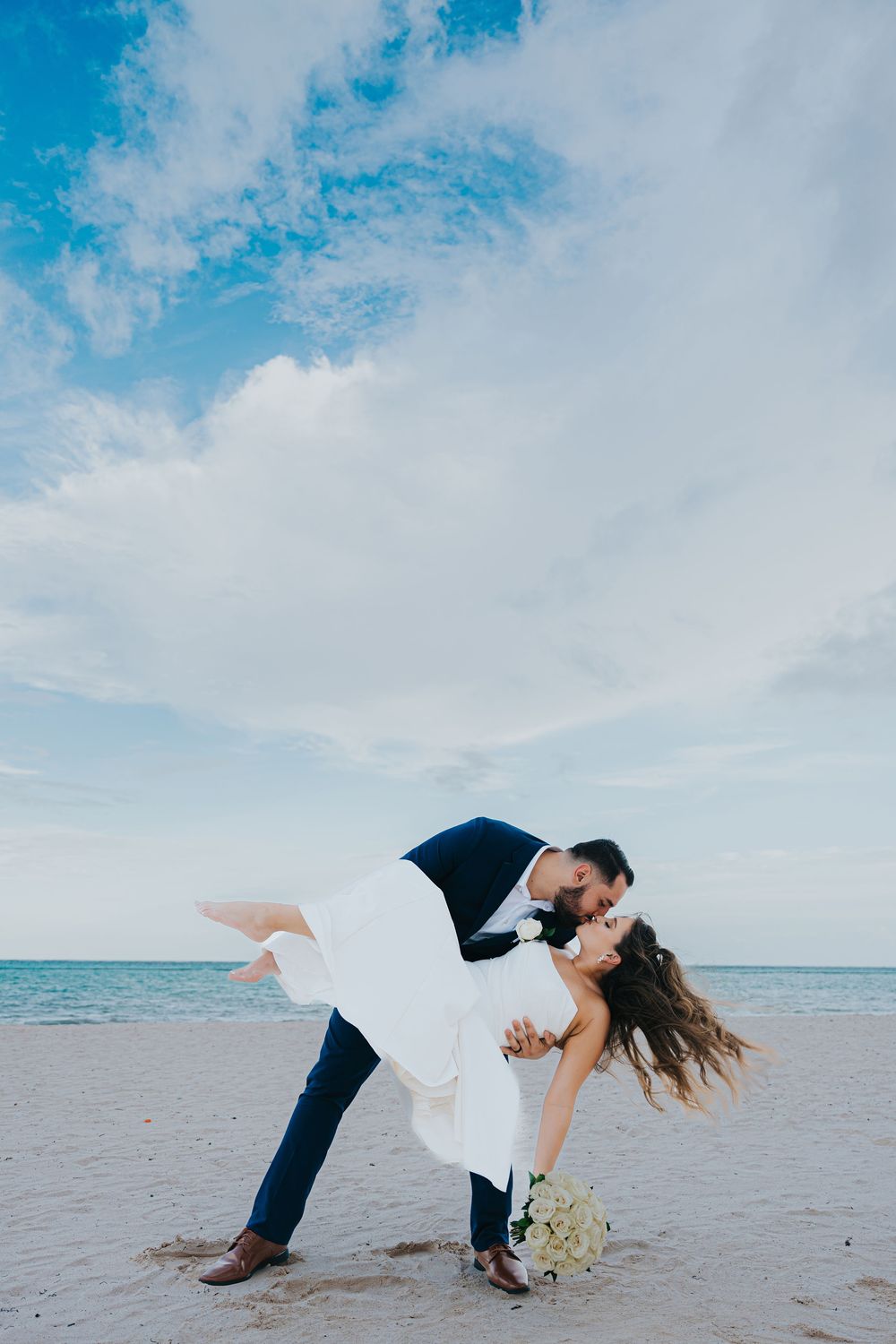 A romantic beach wedding photo captures a dip pose against a serene ocean backdrop with blue skies and wispy clouds.