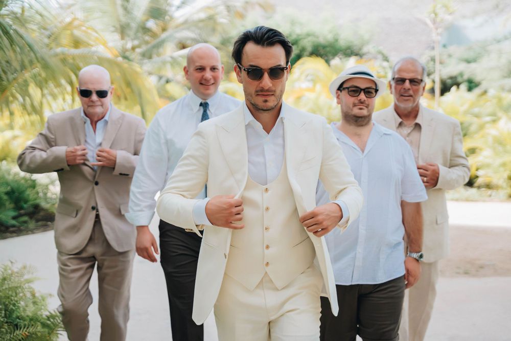 A group of men in light colored suits and sunglasses walk in formation with tropical palm trees in the background.