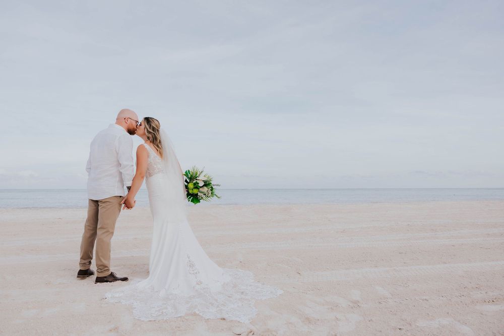 A couple shares a romantic kiss on an empty beach while holding a bouquet against a misty coastal backdrop.