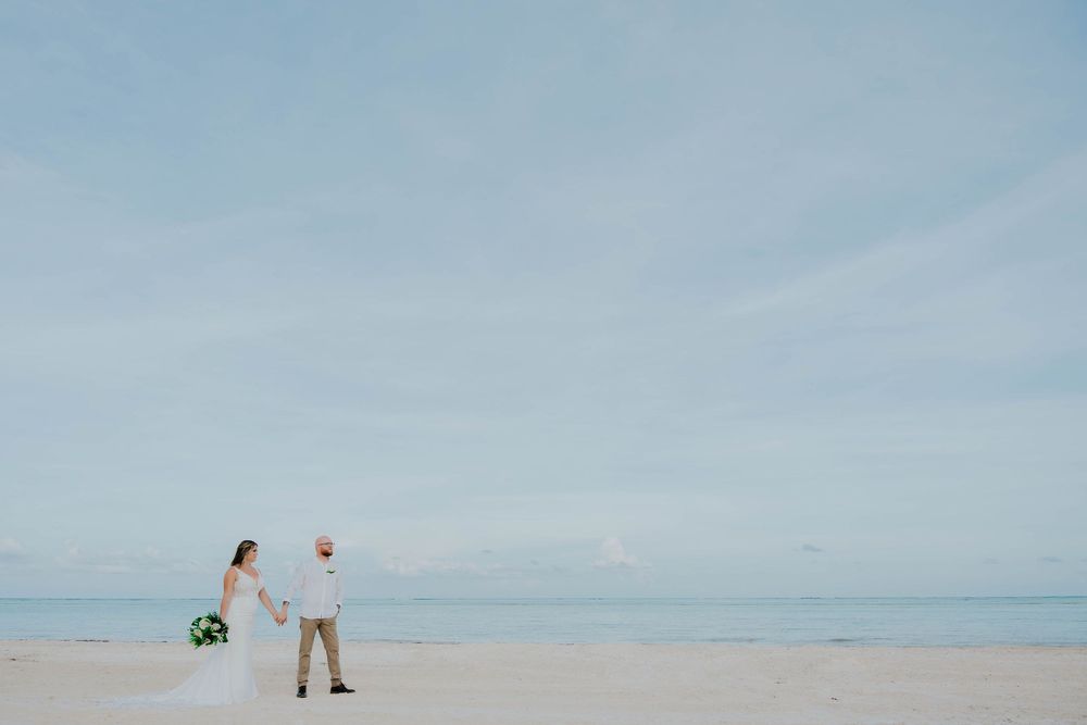 Two figures stand together on an empty beach against a soft blue sky and ocean backdrop.