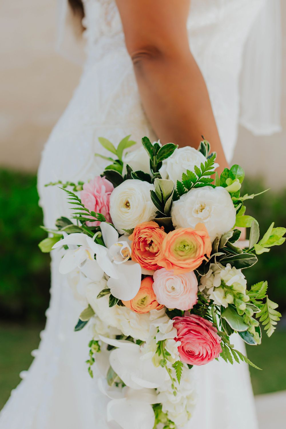 Elegant bridal bouquet featuring white peonies, coral ranunculus, and pink roses with fresh greenery against a white dress.