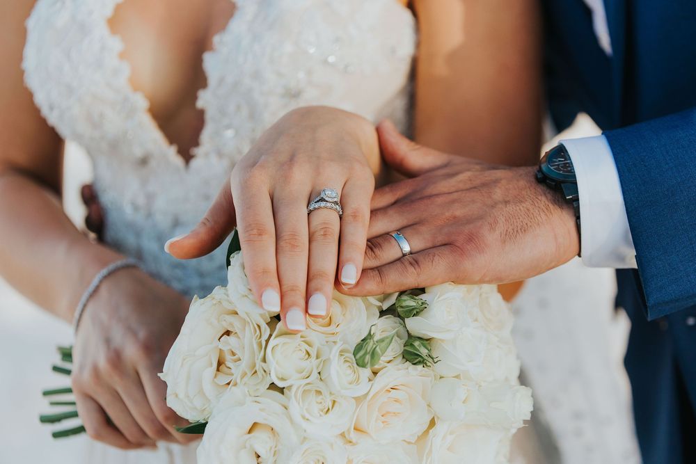 Close-up of newlywed couple's hands with rings resting on white rose bouquet.