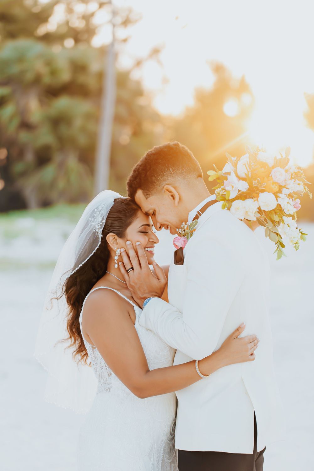 Romantic wedding couple embraces during golden hour with sunlight streaming through flowers in background.