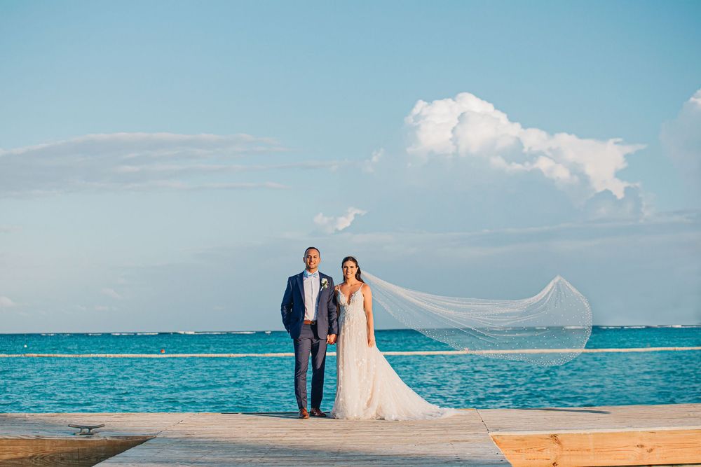 A couple stands on a beach pier with a flowing veil catching the breeze against turquoise ocean and blue sky backdrop.